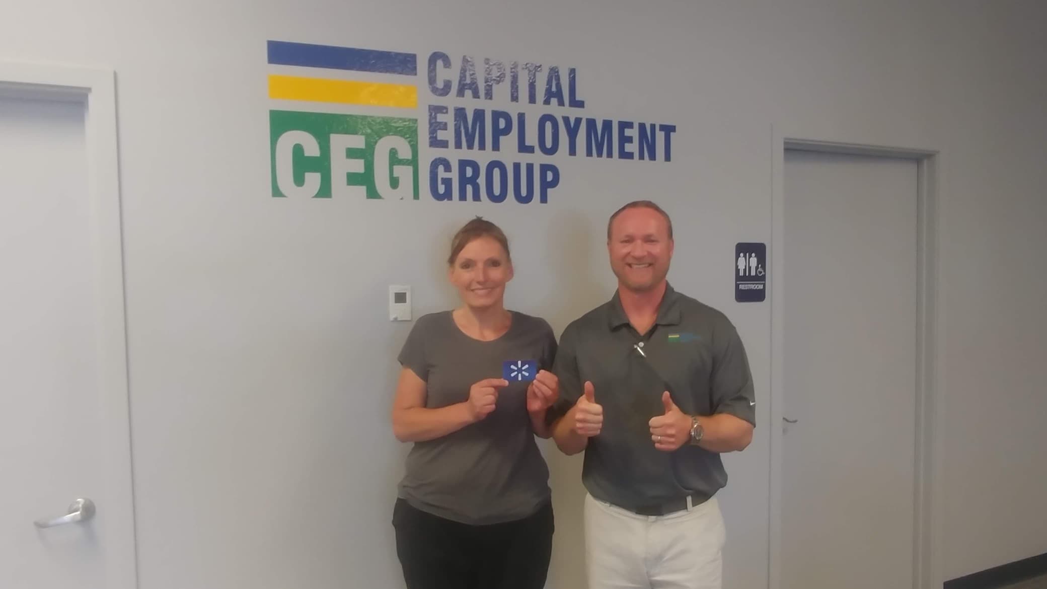 Two individuals pose together in front of a Capital Employment Group sign, smiling and dressed professionally.