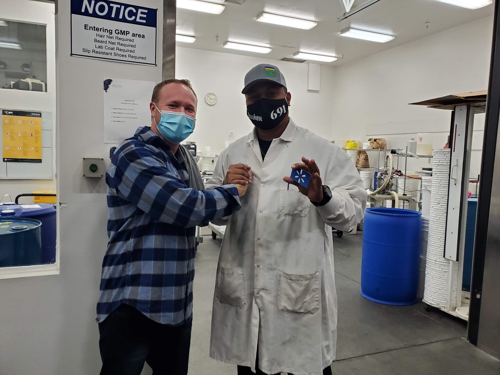 Two men in lab coats and masks stand side by side in a laboratory setting, posing for the camera.