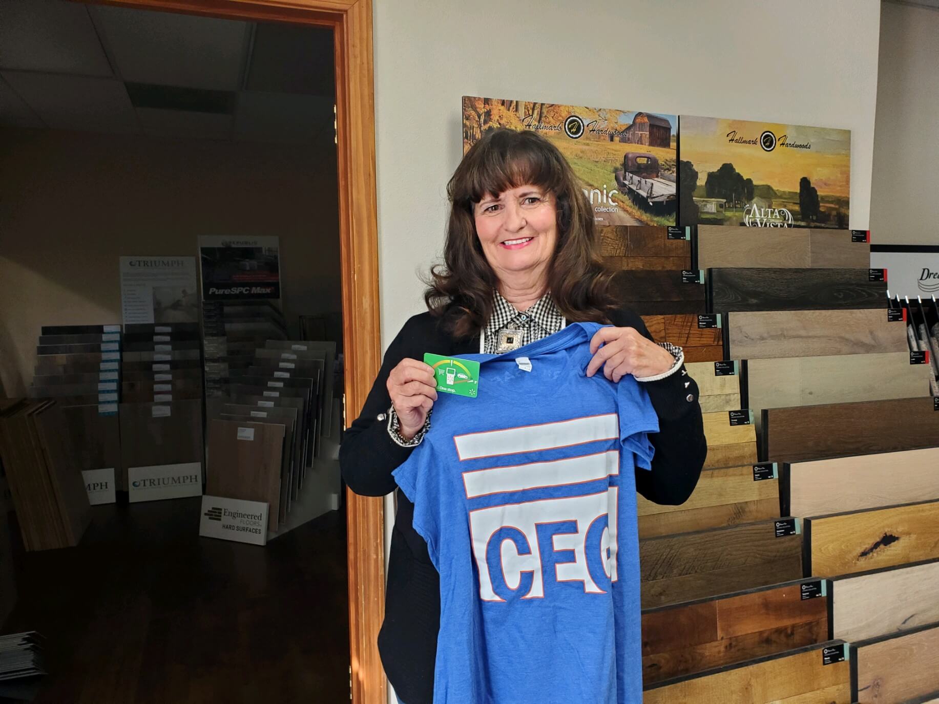 A woman stands in a store, holding a blue shirt while also holding a green card.