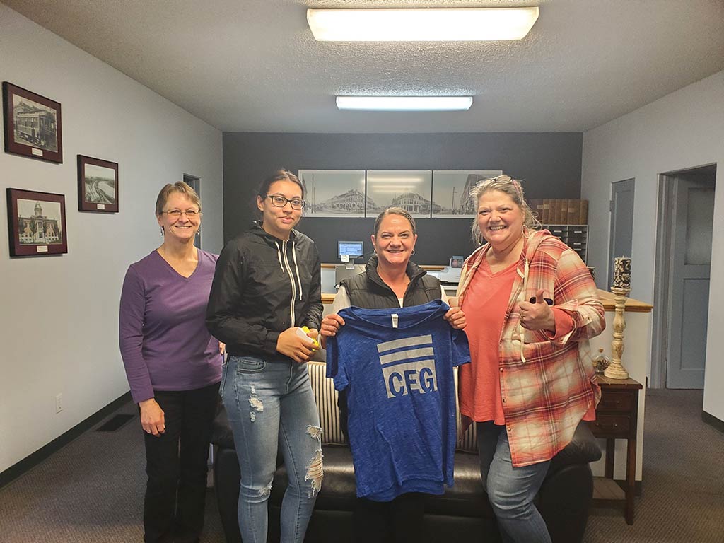Four women in an office hold up a t-shirt, smiling and showcasing the design to the camera.