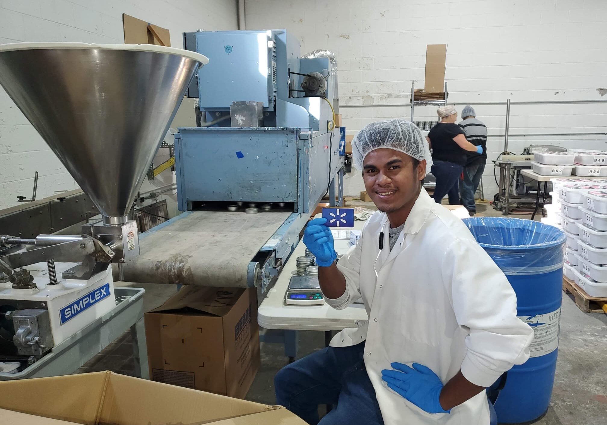 A man wearing a white coat and blue gloves, sitting in a factory setting, appears focused on his work.