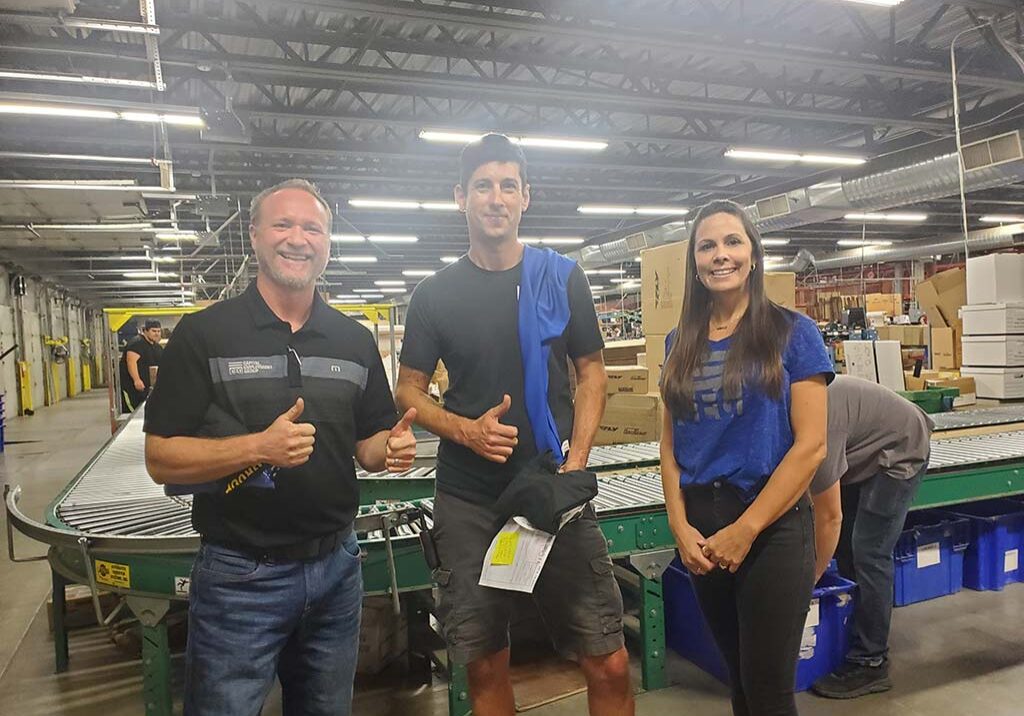 Three individuals in a factory, with a man and woman smiling and giving thumbs up, showcasing teamwork and positivity.